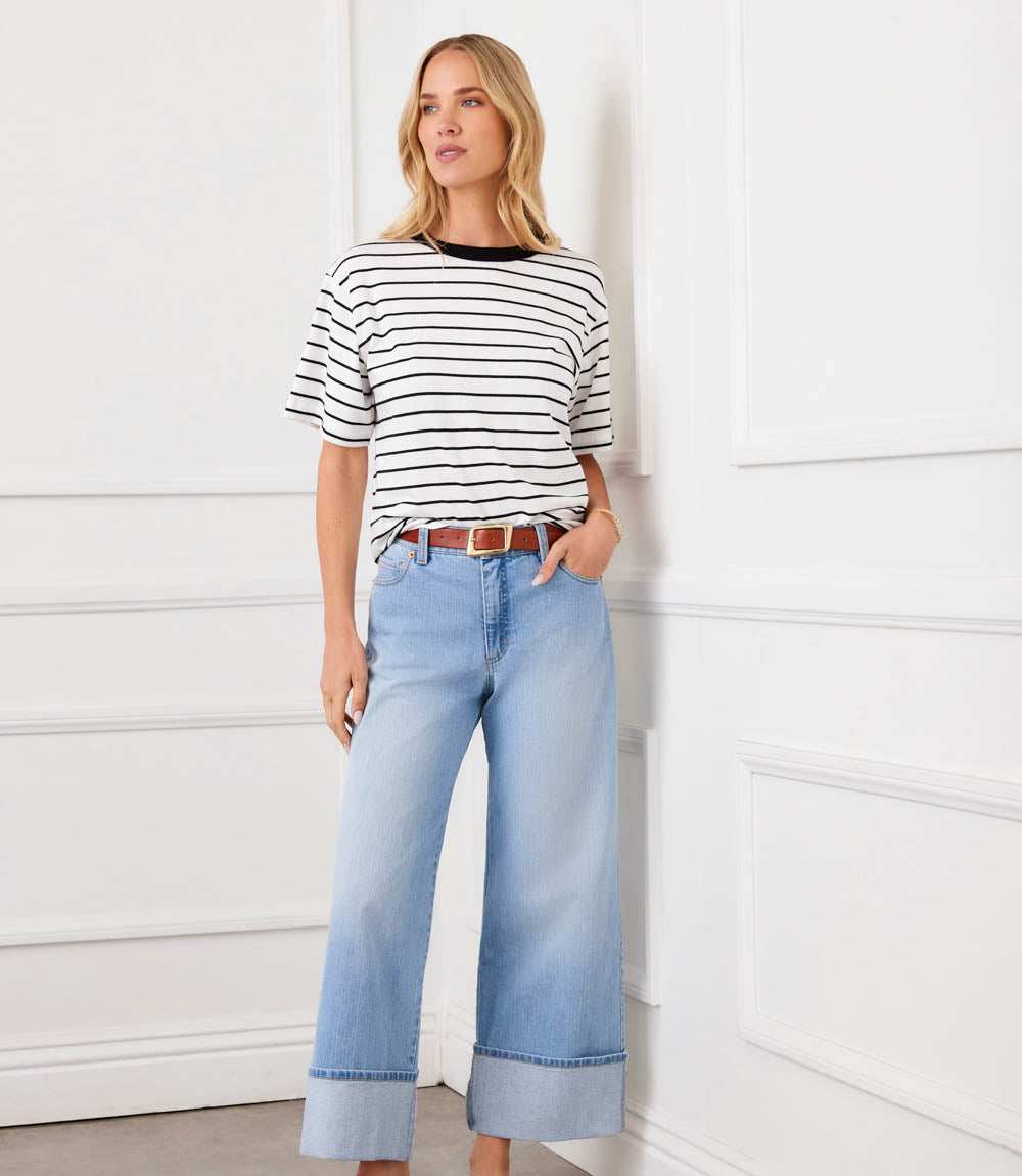 A woman stands against a white paneled wall wearing a black and white horizontally striped short-sleeve top, light-wash blue wide-leg cropped jeans, and a brown belt.