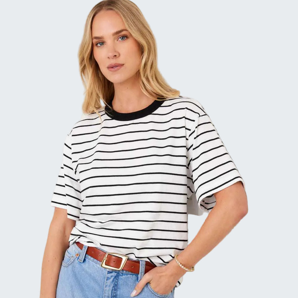 A woman wearing a black and white horizontally striped short-sleeve t-shirt with a black crew neck, tucked into light blue jeans with a brown belt, posing against a white paneled wall.
