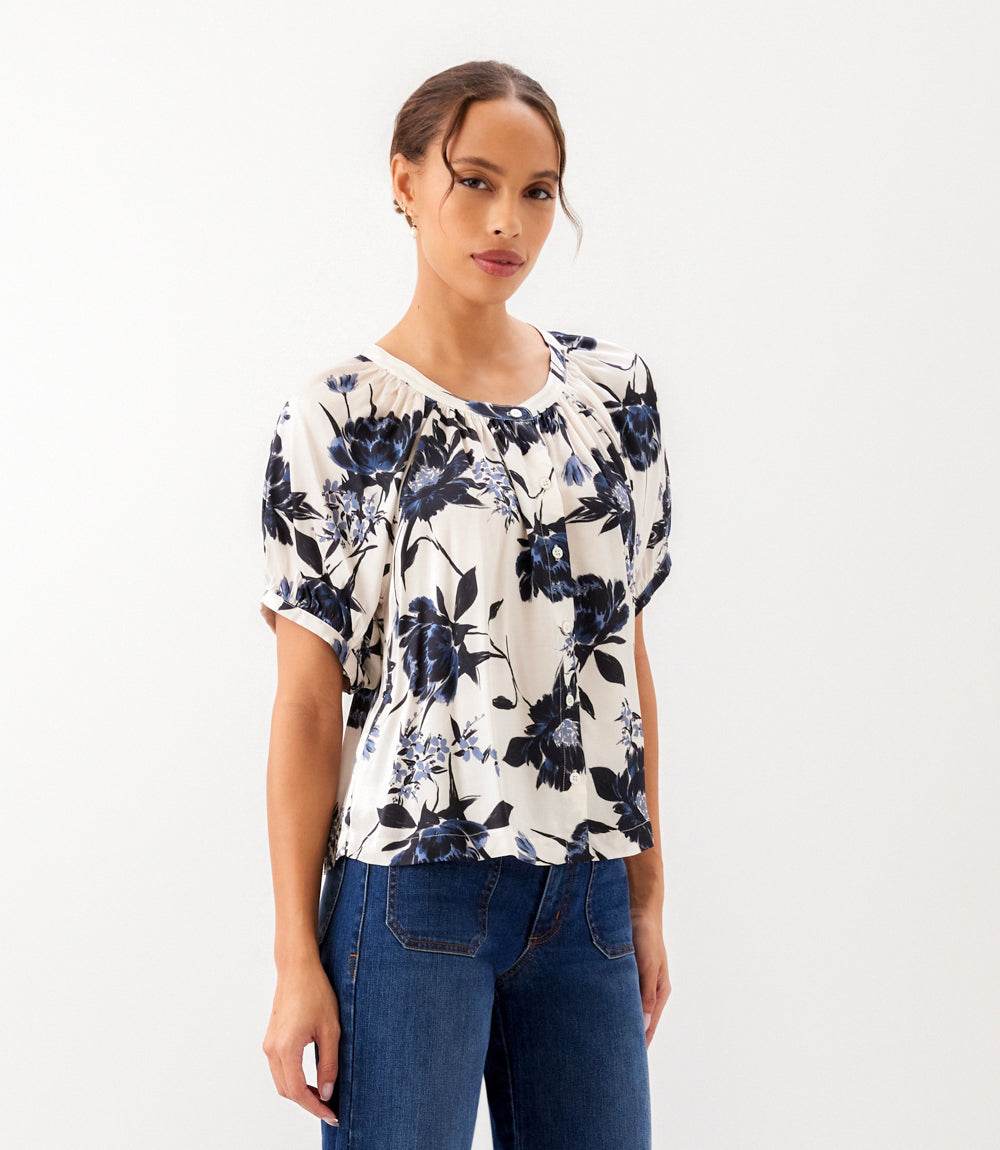 A woman modeling a short-sleeve blouse with a navy blue floral print on an off-white background, paired with blue jeans, against a plain white studio background.