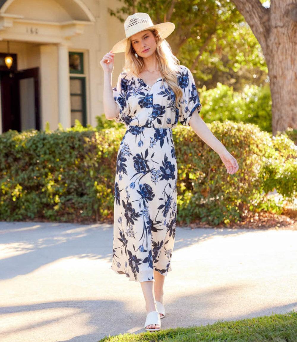 A woman wearing a white midi dress with a navy blue floral print, a cream straw hat, and white slide sandals walks along a sunlit paved path in front of a house and green hedges.