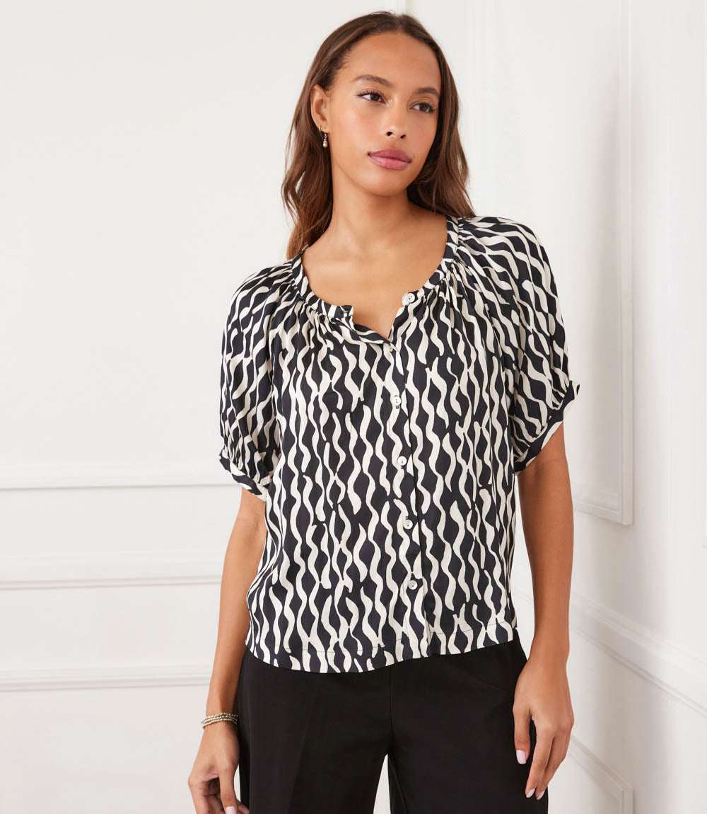 A woman modeling a short-sleeve blouse with a black and white wavy abstract pattern, paired with black trousers, standing against a white paneled wall.