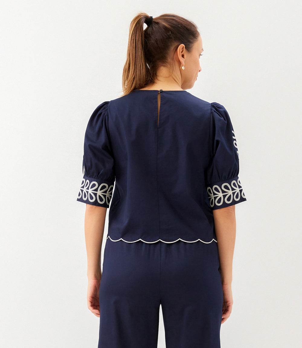 Woman wearing a navy blue top with white embroidery on sleeves and hemline against a white background