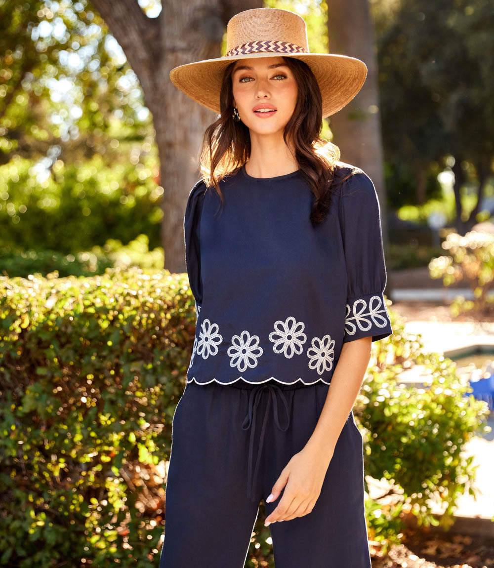 A woman wearing a navy blue two-piece set with white embroidered scalloped flower details, a matching wide-brim straw hat, and a drawstring waist, standing outdoors in front of green bushes and trees.