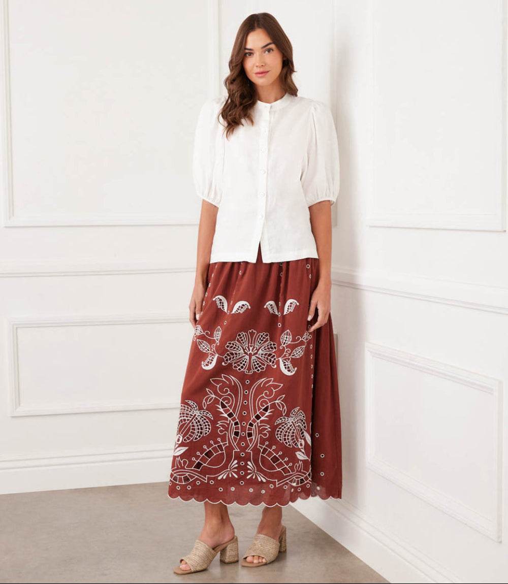 Woman wearing a white blouse and brown patterned skirt against a white wall.