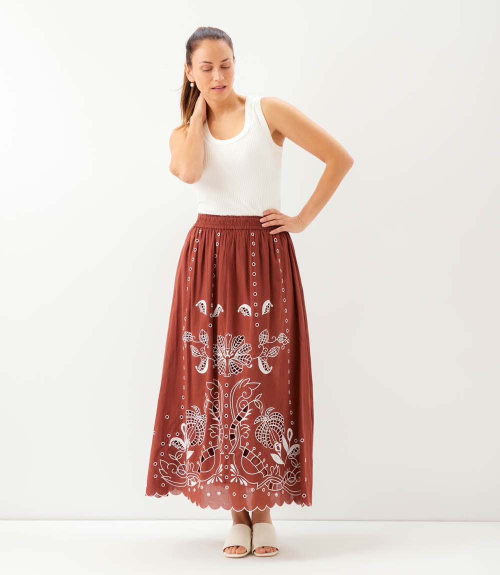 A woman modeling a rust-brown maxi skirt with white floral embroidery and a scalloped hem, paired with a white tank top and beige flat sandals, against a plain white studio background.