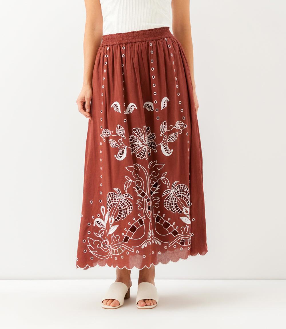 A rust-colored midi skirt with white floral embroidery, shown on a model wearing beige slide sandals against a plain white studio background.