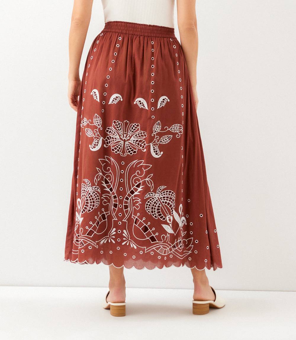 A woman wearing a rust-brown midi skirt with white floral embroidery and scalloped edges, shown from the back against a plain white studio background.