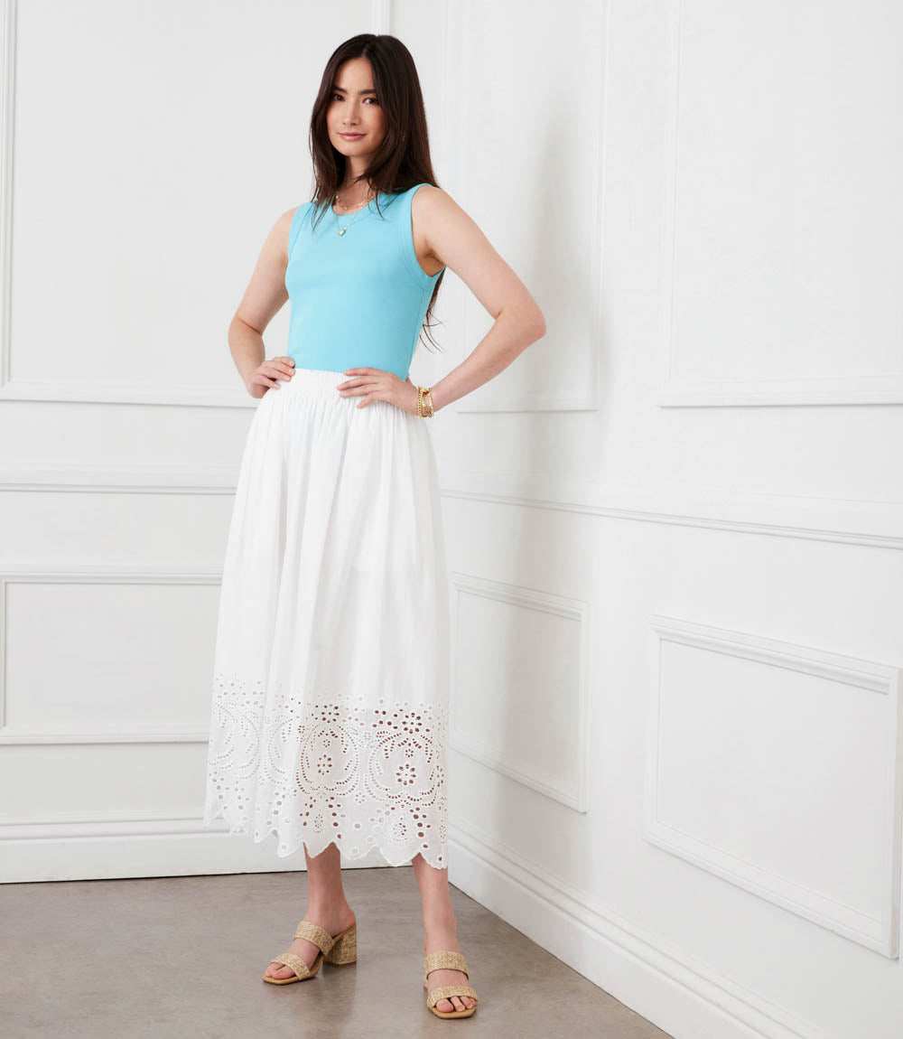 Woman wearing a light blue sleeveless top and white lace skirt against a white wall.