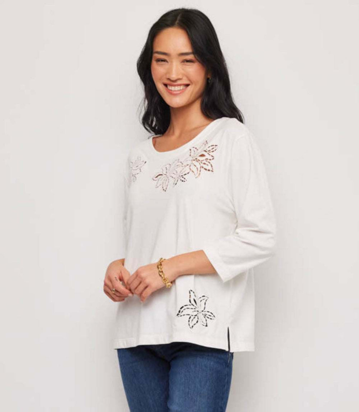 A woman wearing a white three-quarter sleeve top with floral embroidery, paired with blue jeans, standing against a plain white background.