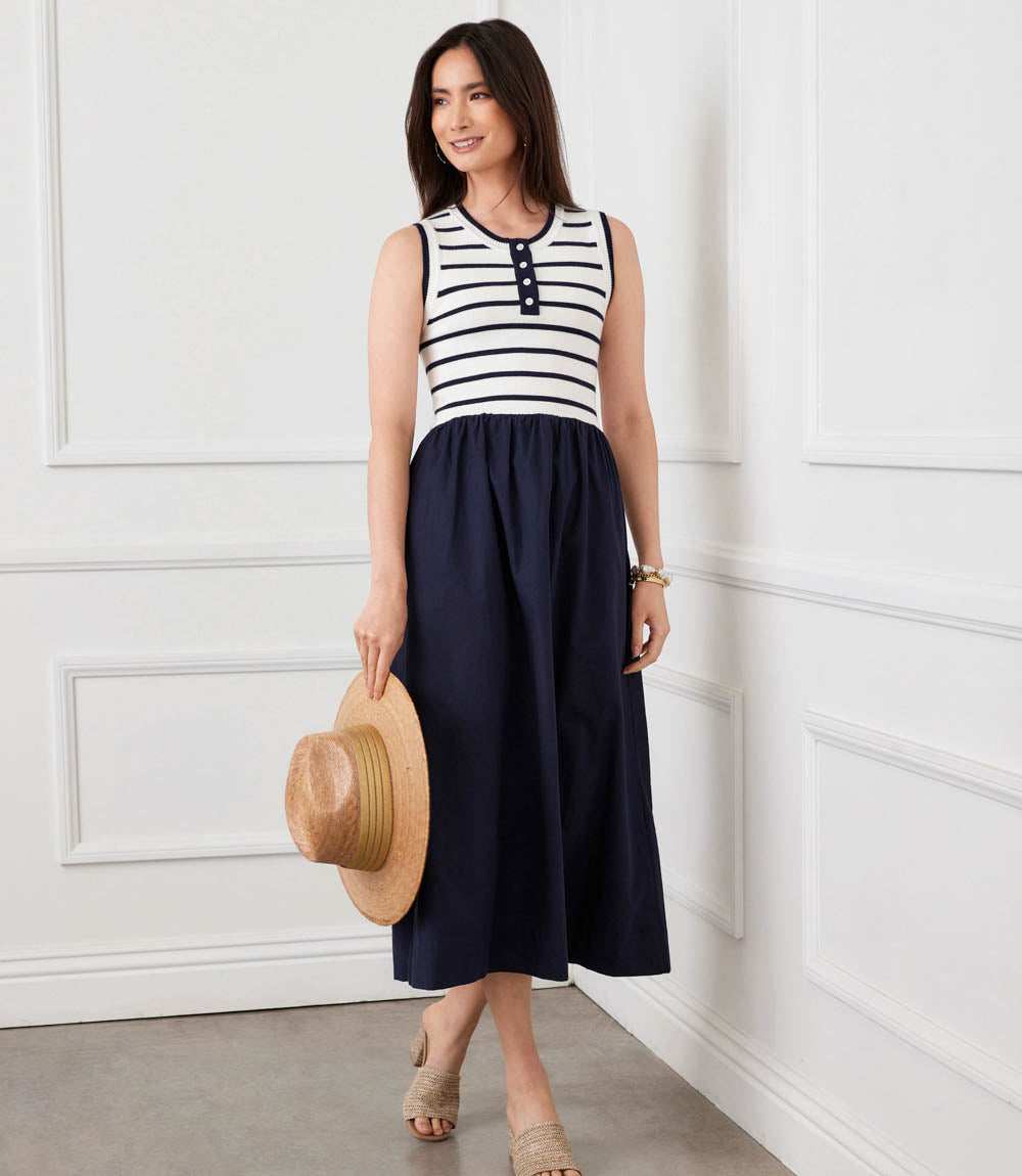 Woman wearing a black and white striped sleeveless top and navy skirt, holding a straw hat against a white paneled wall.