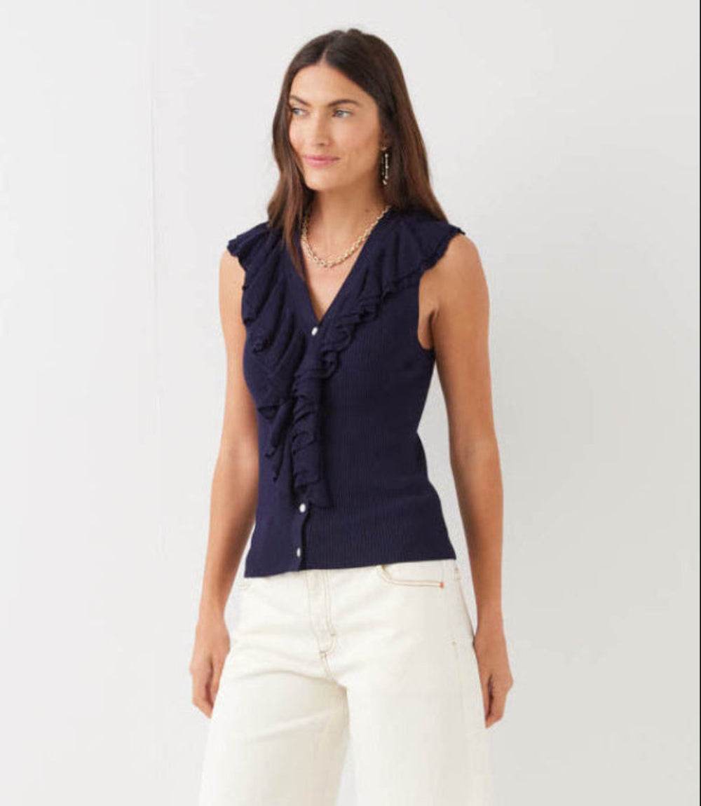 Woman wearing a navy blue ruffled top and white pants on a light gray background