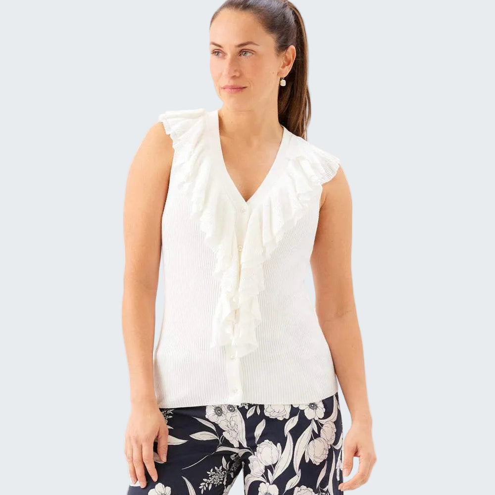 A woman wearing a white sleeveless knit top with layered ruffles down the front, paired with navy floral pants, against a plain white studio background.