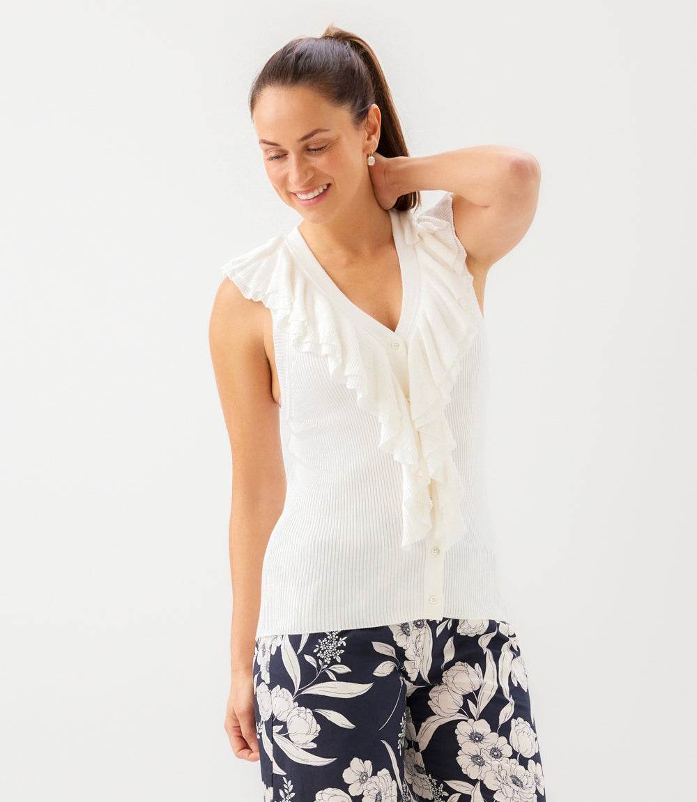 A woman wearing a white sleeveless ribbed knit top with ruffle trim and dark floral print pants, posing against a plain white studio background.