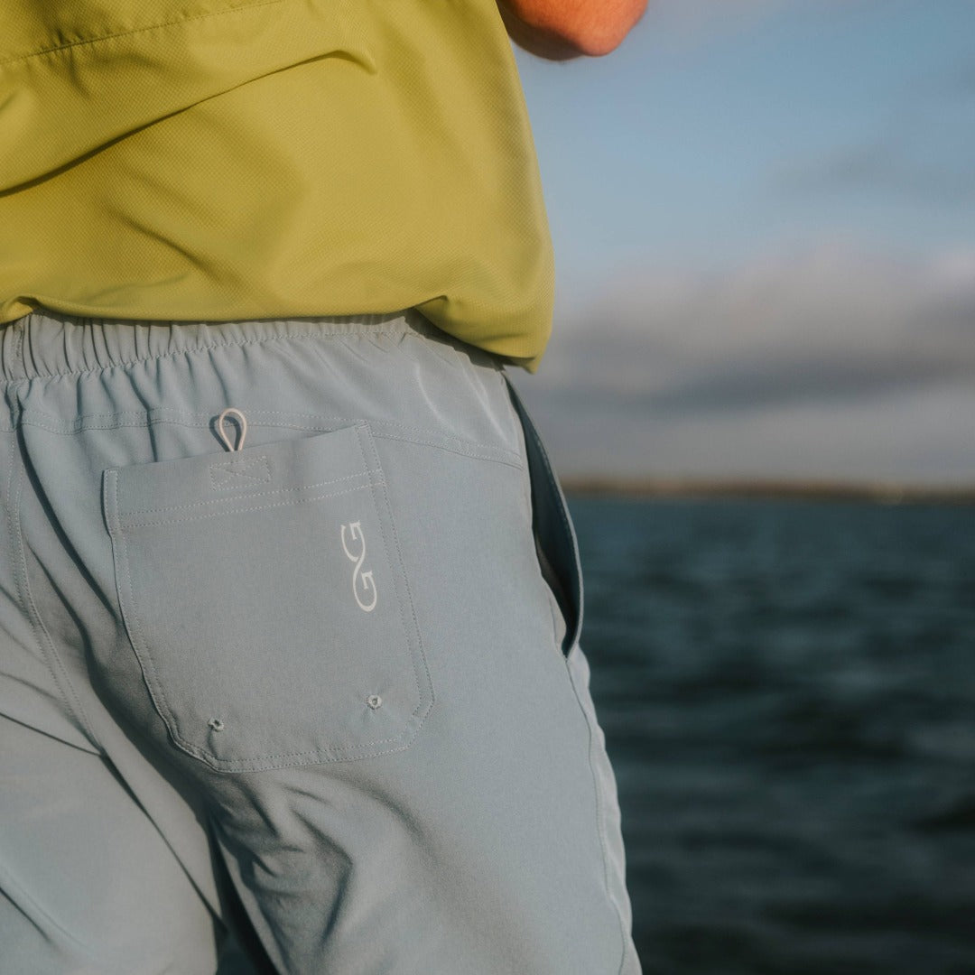 Person wearing a green jacket and gray pants with a logo, standing by a body of water.