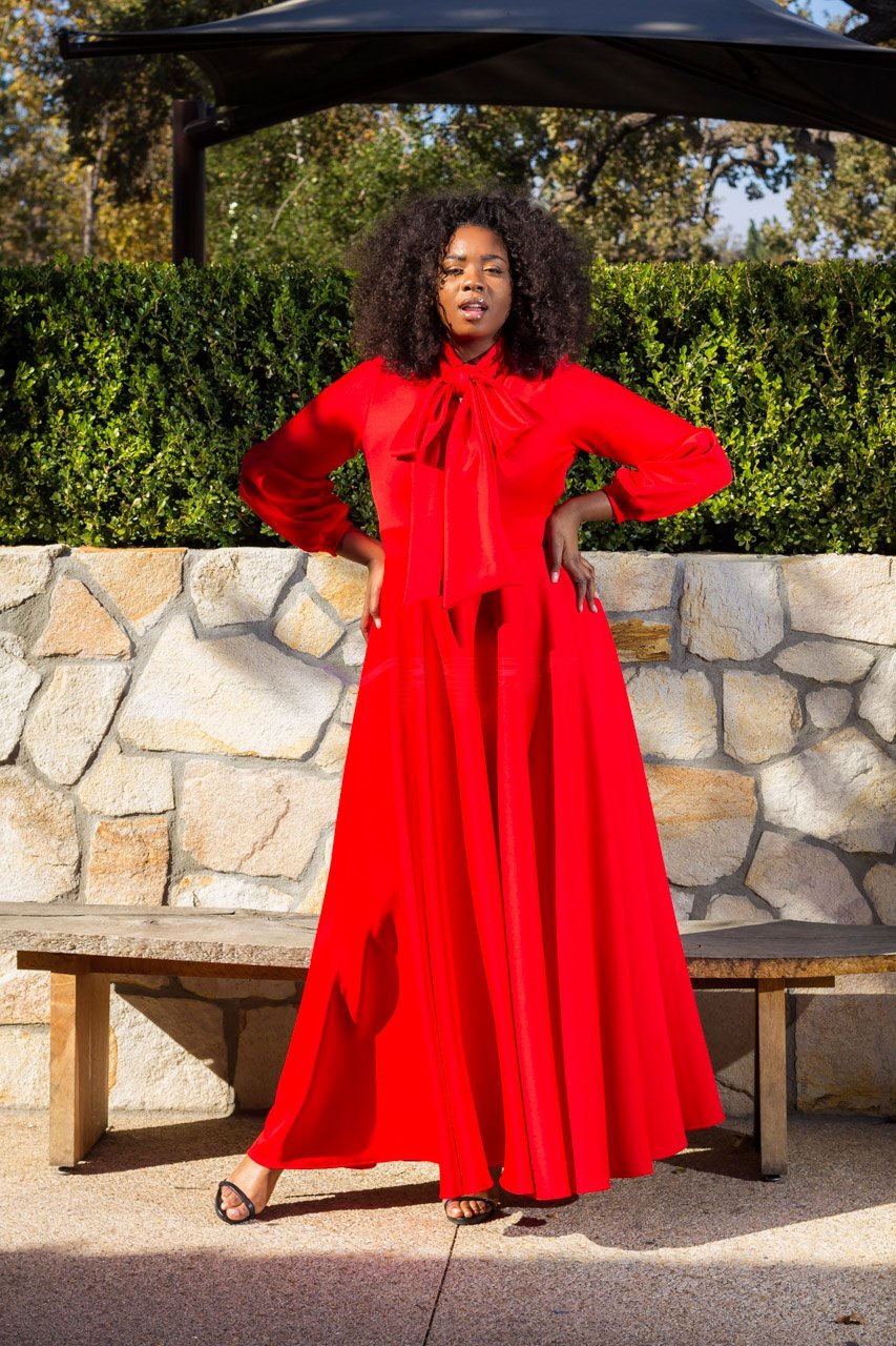 A woman wearing a bright red long-sleeve maxi dress with a large bow tie neckline stands outdoors in front of a stone wall and hedge, with a bench and patio umbrella visible.