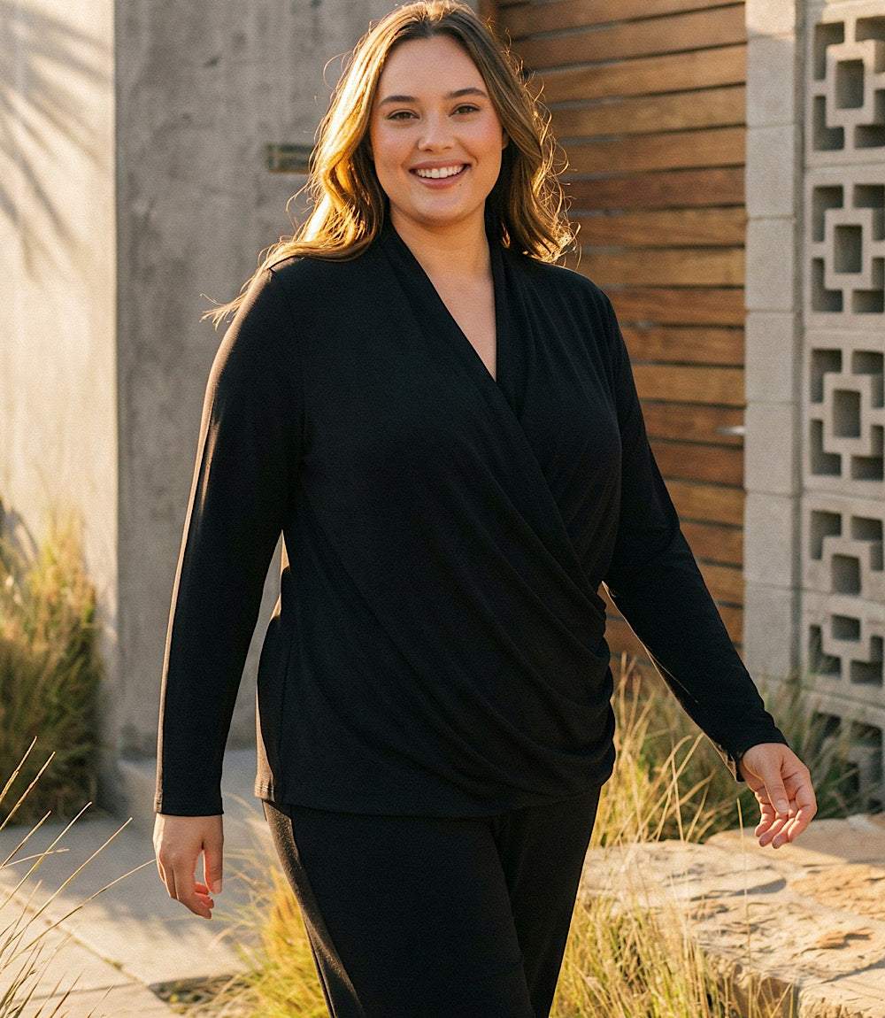 A woman wearing a black long-sleeve wrap-style top and matching black pants, standing outdoors in front of a modern building with concrete and wood panels.