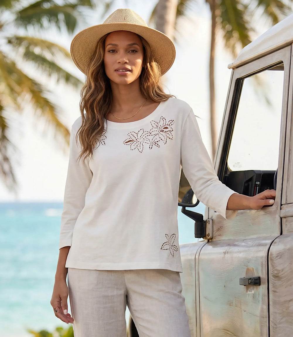 Woman in a white outfit with floral embroidery standing next to a vehicle by the beach.