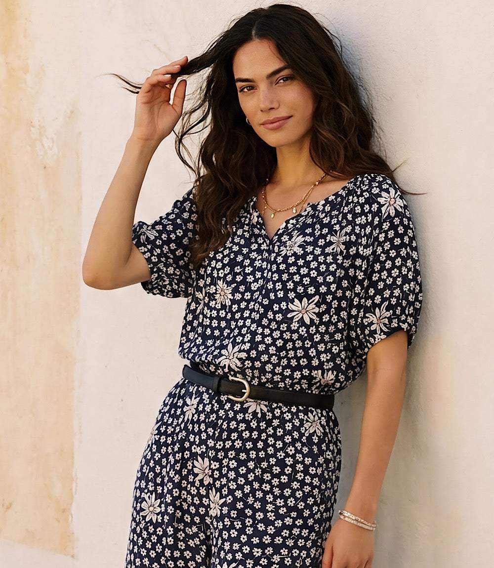 A woman with long dark wavy hair leans against a textured off-white wall, wearing a navy blue short-sleeve jumpsuit with a white floral print and a black belt.