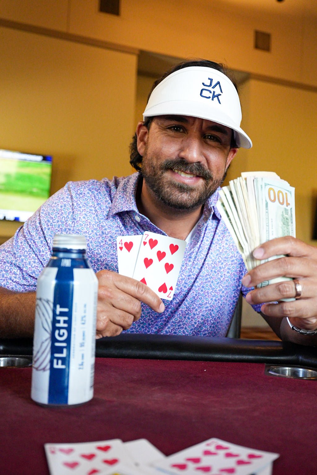 Man holding playing cards and cash at a poker table with a bottle of 'Flight' drink.