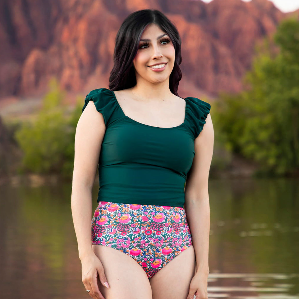 Woman in a green top and colorful floral swimsuit standing by a body of water with mountains in the background.