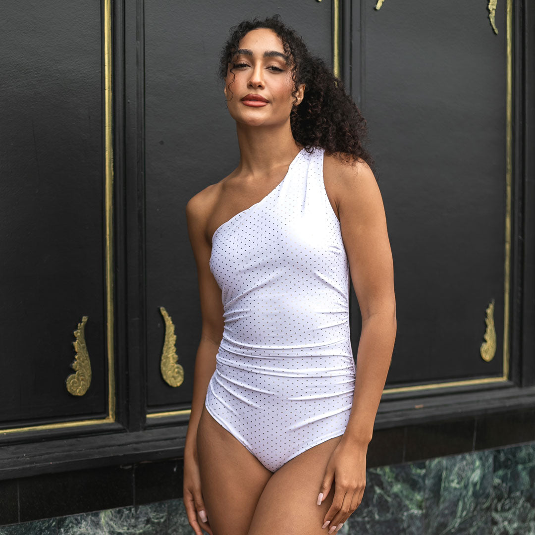 Woman wearing a white polka dot swimsuit standing in front of a black door with gold accents.