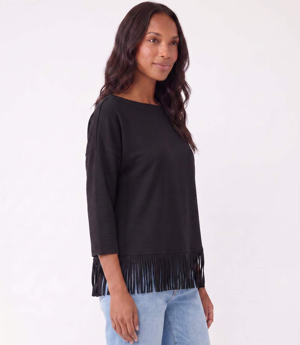 A woman wearing a black three-quarter sleeve top with fringe trim at the hem, paired with light blue jeans, photographed against a plain white studio background.