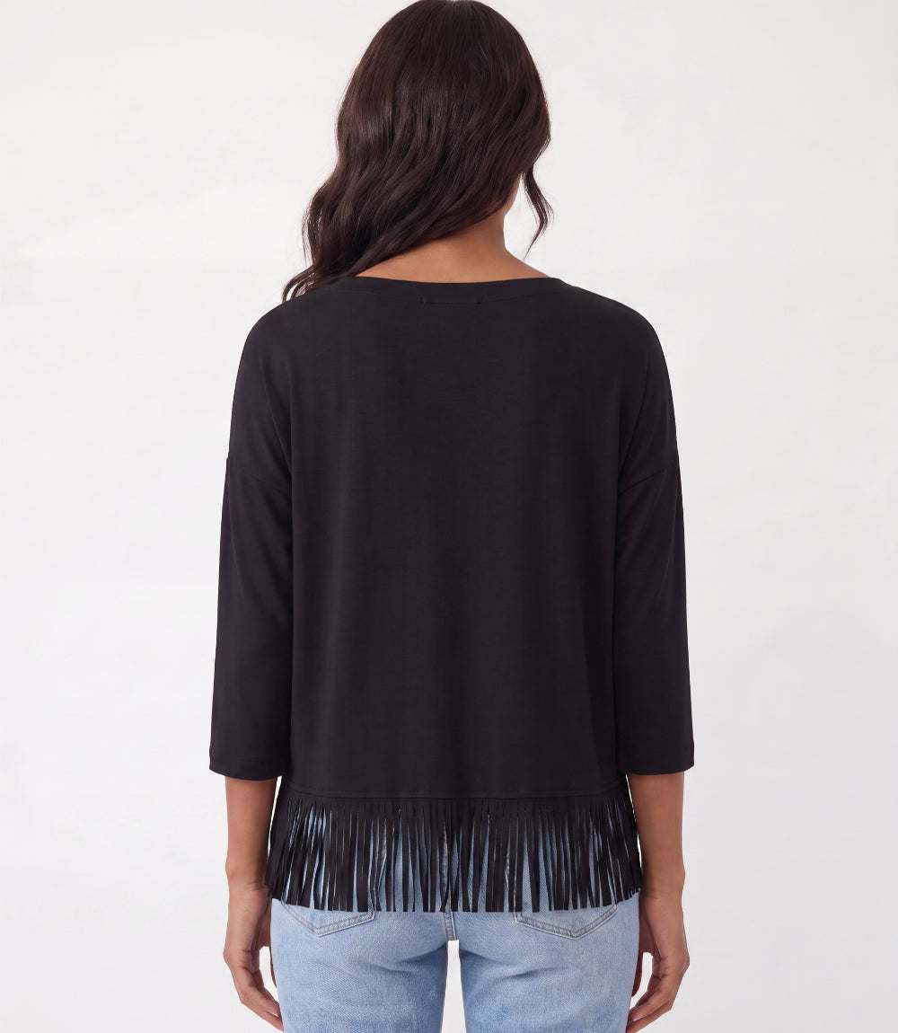 A woman viewed from the back wearing a black three-quarter sleeve top with fringe trim at the hem, paired with light blue jeans, against a plain white background.