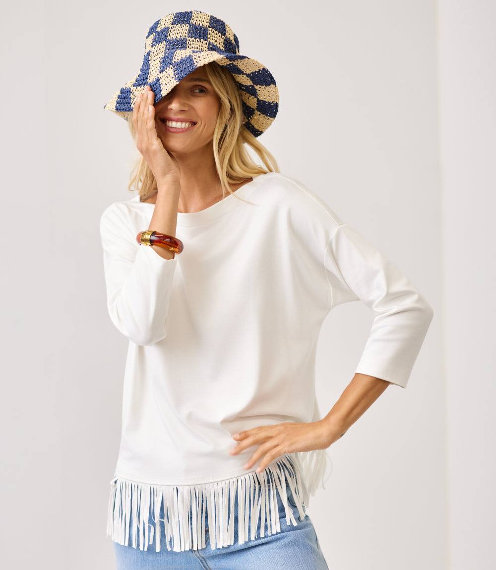 A woman wearing a white long-sleeve top with fringe trim at the hem, paired with a blue and beige checkered bucket hat and light blue jeans, posing against a light gray studio background.