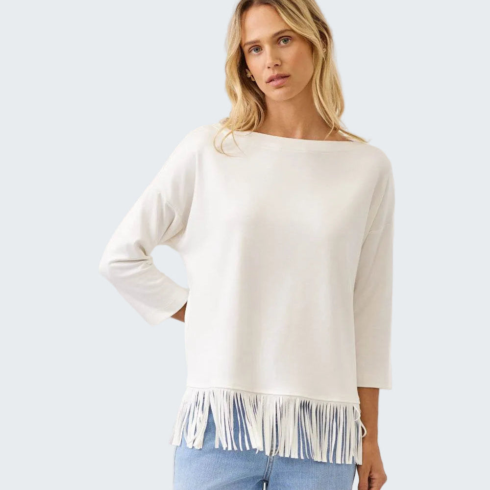 A woman modeling a cream-colored knit sweater with a wide boat neckline and fringe trim along the hem, paired with light blue jeans against a plain white studio background.