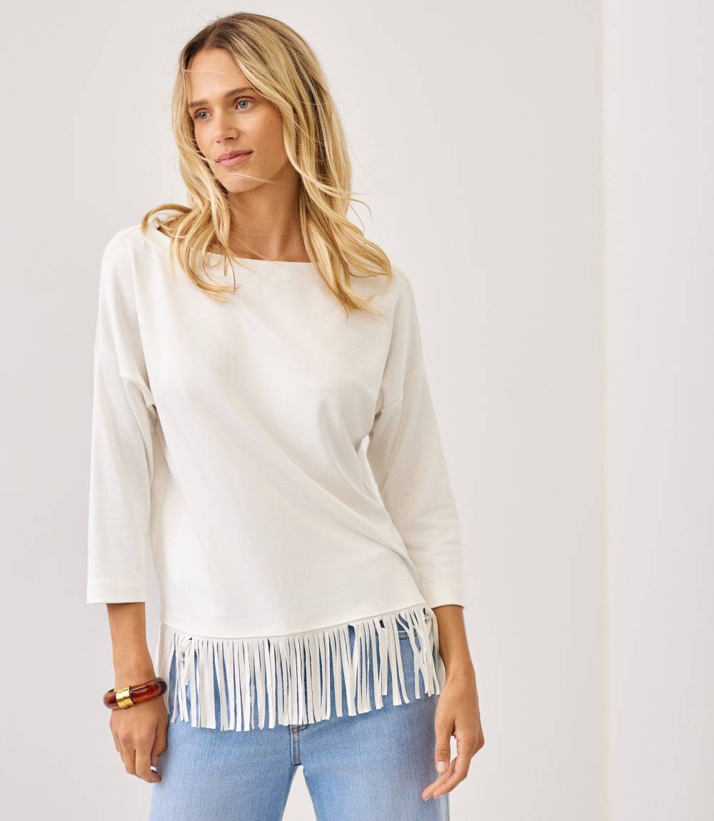 A woman modeling a cream-colored three-quarter sleeve top with a fringe hem, paired with light blue jeans, against a plain off-white background.