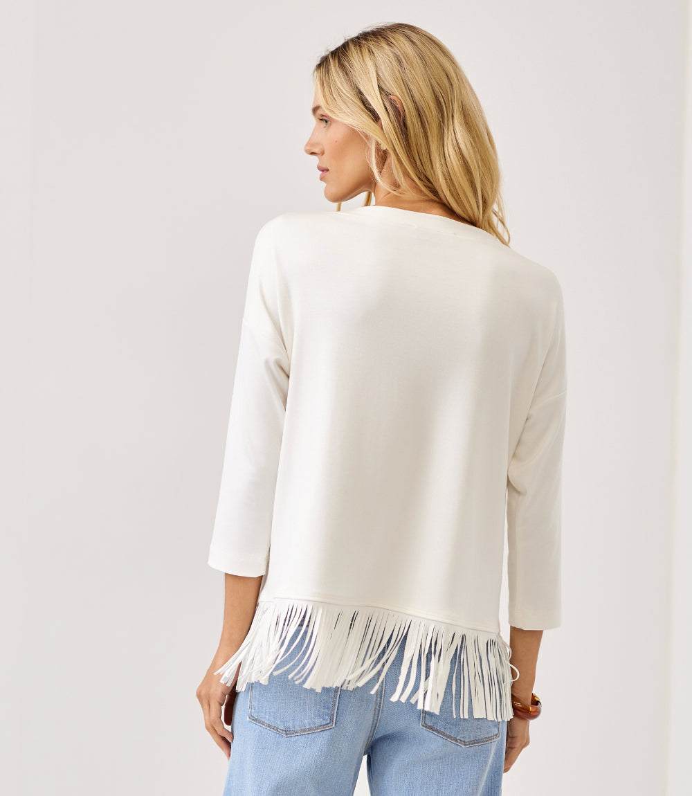 A woman shown from the back wearing a cream-colored knit top with fringe trim at the hem, paired with light blue jeans, against a plain off-white background.