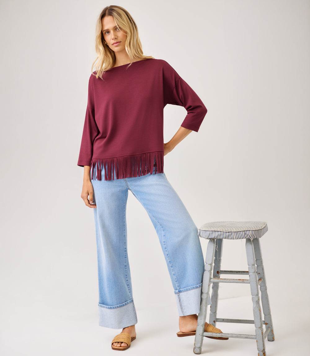 A woman models a burgundy long-sleeve top with fringe trim, paired with light blue wide-leg jeans and brown slide sandals, standing next to a rustic wooden stool against a plain light gray background.