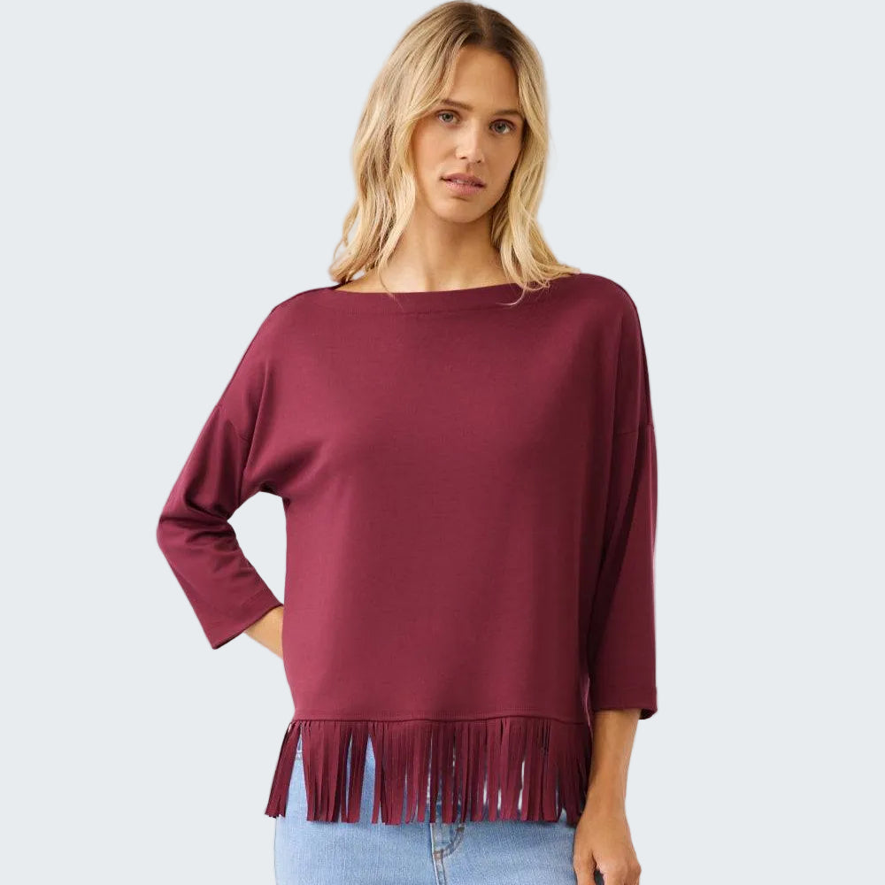 A woman wearing a burgundy long-sleeve top with fringe trim at the hem, paired with light blue jeans, against a plain light gray studio background.