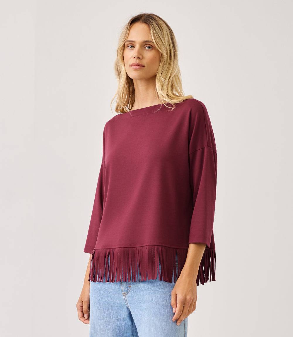 A woman wearing a loose-fitting, three-quarter sleeve top in deep burgundy with fringe trim along the hem, paired with light blue jeans, against a plain light gray studio background.