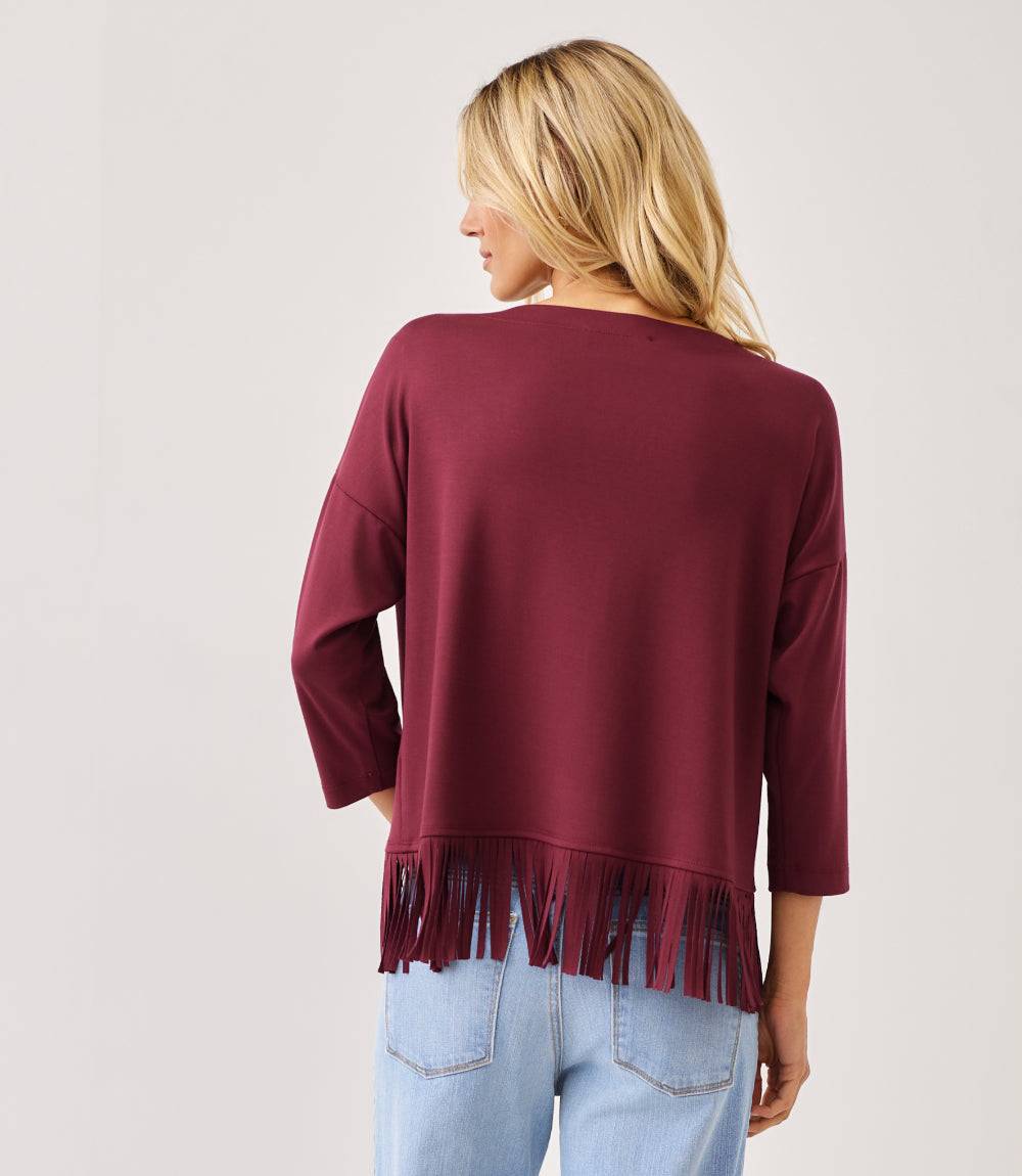 A woman shown from the back wearing a burgundy long-sleeve top with fringe trim at the hem, paired with light blue jeans, against a plain light gray studio background.