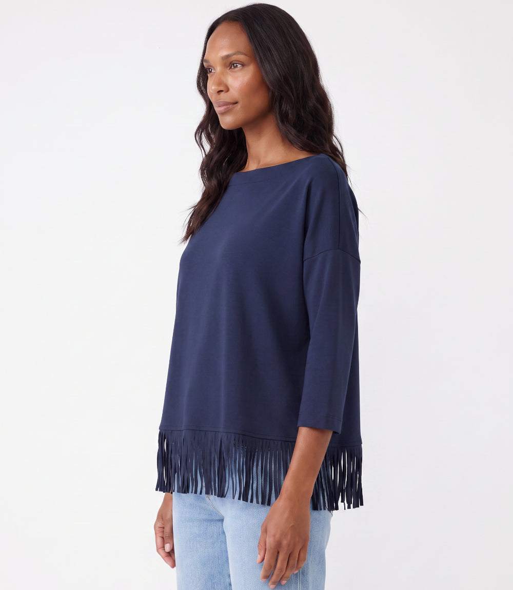 A woman wearing a navy blue boat-neck top with fringe trim along the hem, paired with light blue jeans, photographed against a plain white studio background.