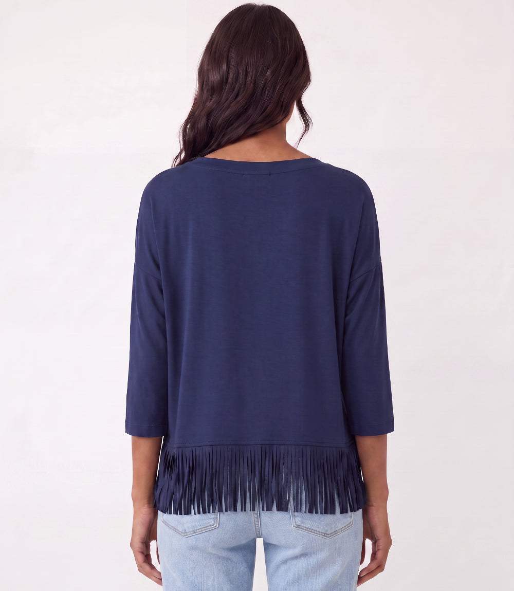 A woman viewed from the back wearing a navy blue three-quarter sleeve top with fringe trim at the hem, paired with light blue jeans, against a plain white background.