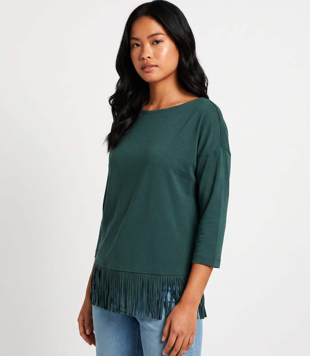 A woman modeling a dark green three-quarter sleeve top with fringe trim along the hem, paired with light blue jeans against a plain white studio background.