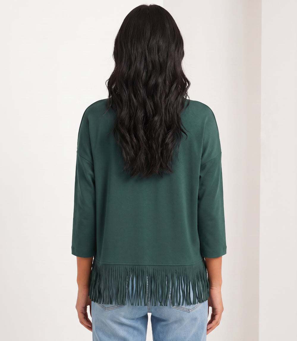 A woman viewed from the back wearing a dark green long-sleeve top with fringe trim at the hem, paired with light blue jeans, against a plain off-white background.