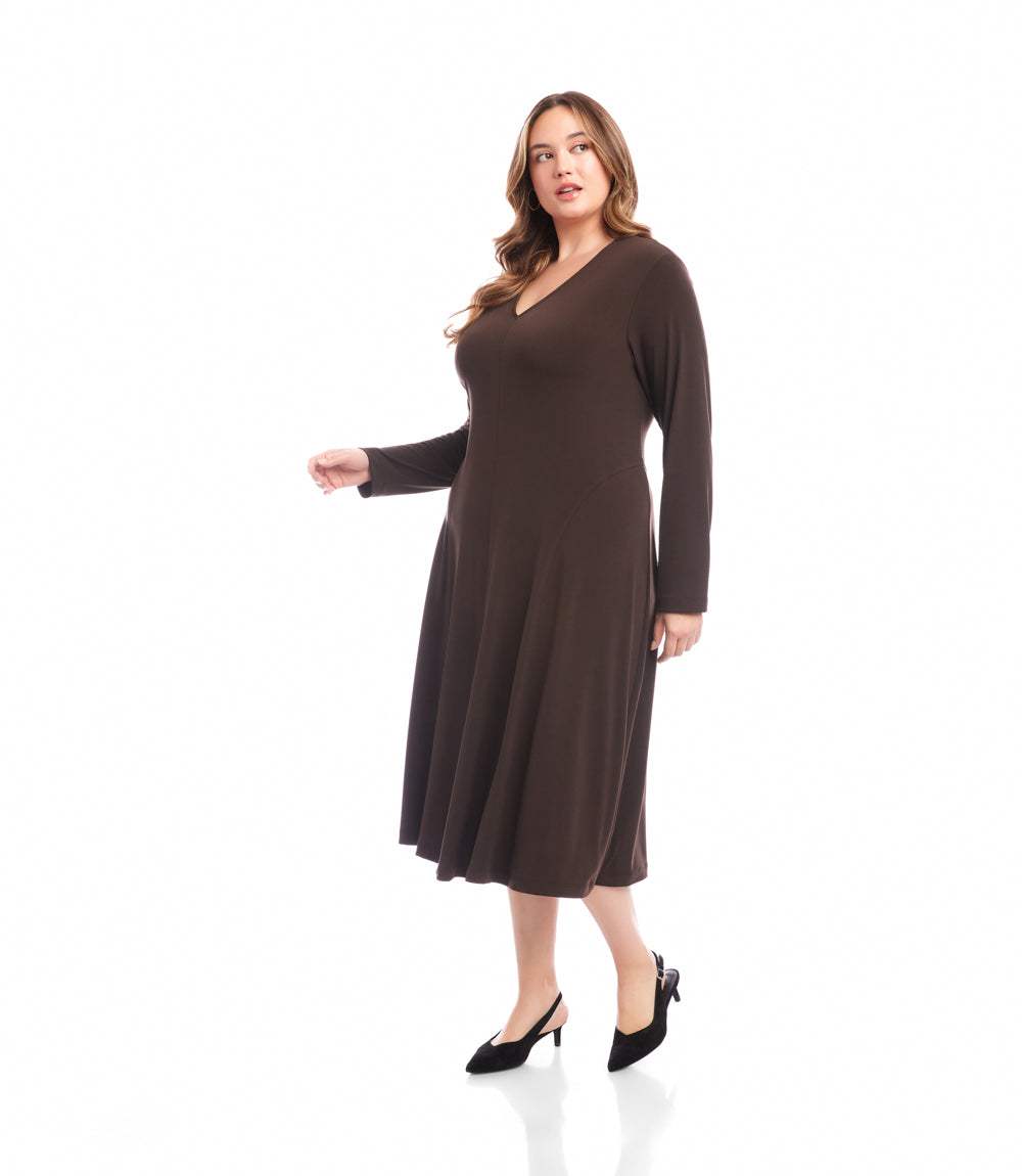 A woman modeling a dark brown long-sleeve midi dress with a V-neckline, paired with black pointed-toe heels, against a solid white studio background.