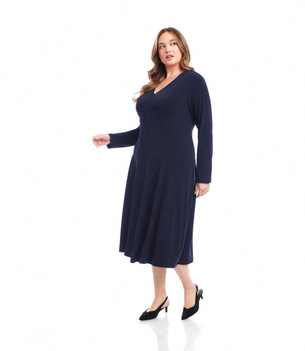 A woman modeling a long-sleeve, V-neck midi dress in solid black, paired with black pointed-toe heels, against a plain white studio background.