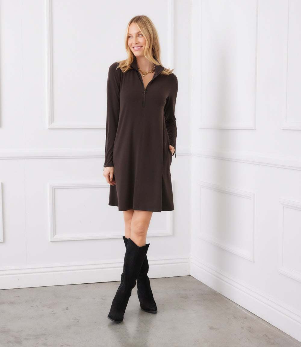 A woman modeling a dark brown long-sleeve shift dress with a half-zip neckline, paired with black ankle boots, standing in a white paneled room.
