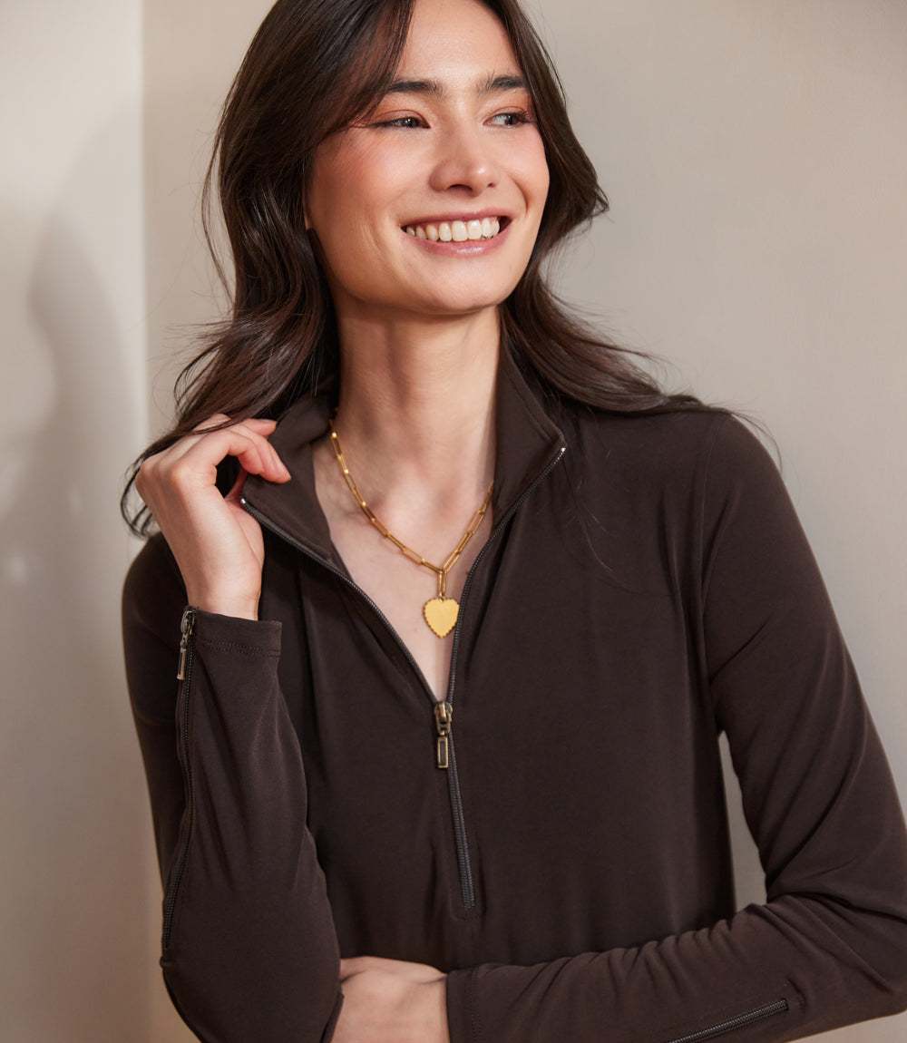 A woman wearing a dark brown quarter-zip long-sleeve top with a gold heart pendant necklace, posing against a neutral beige background.