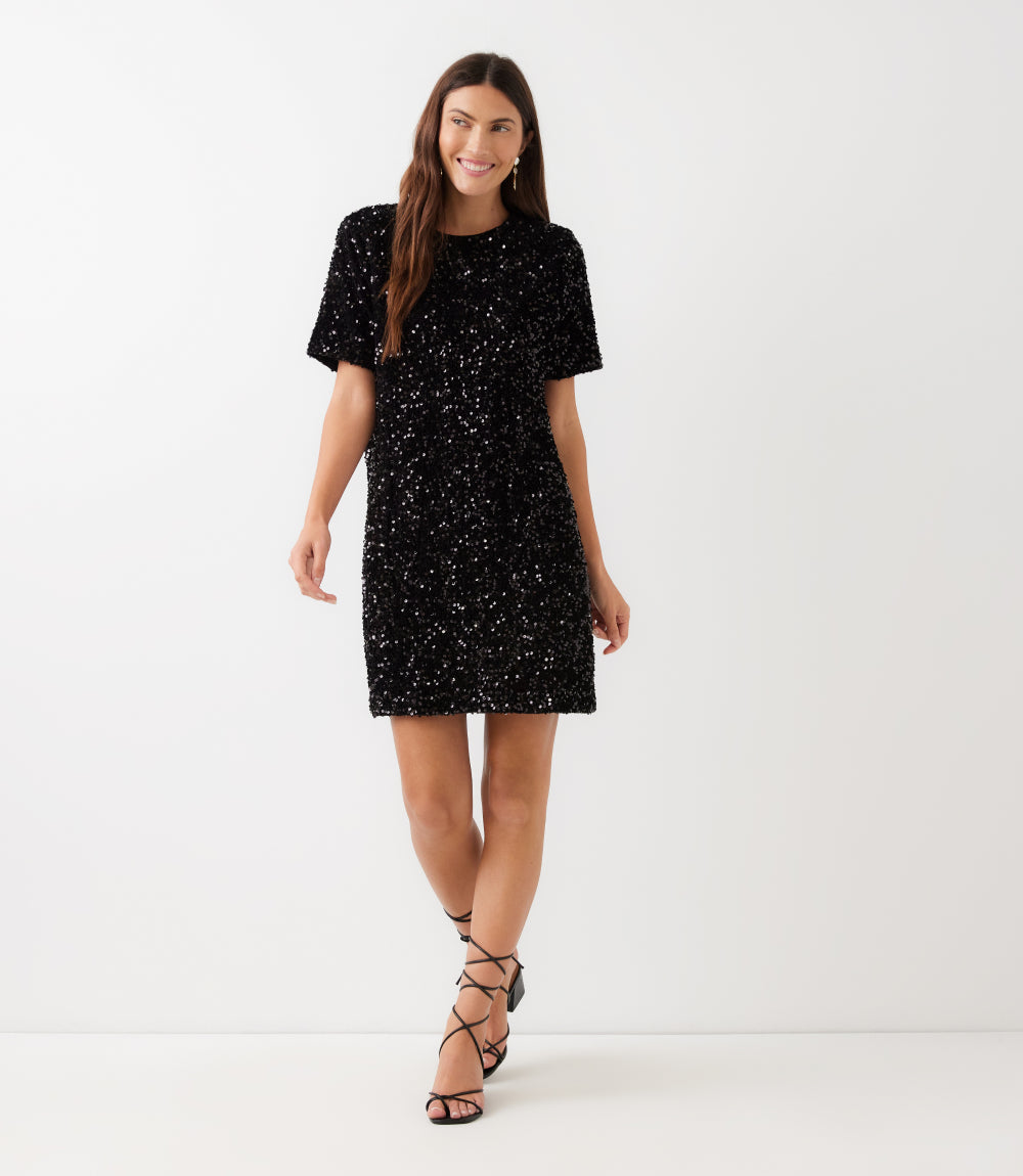A woman modeling a short-sleeve black sequined mini dress with a round neckline, paired with black lace-up strappy heels, against a plain white studio background.