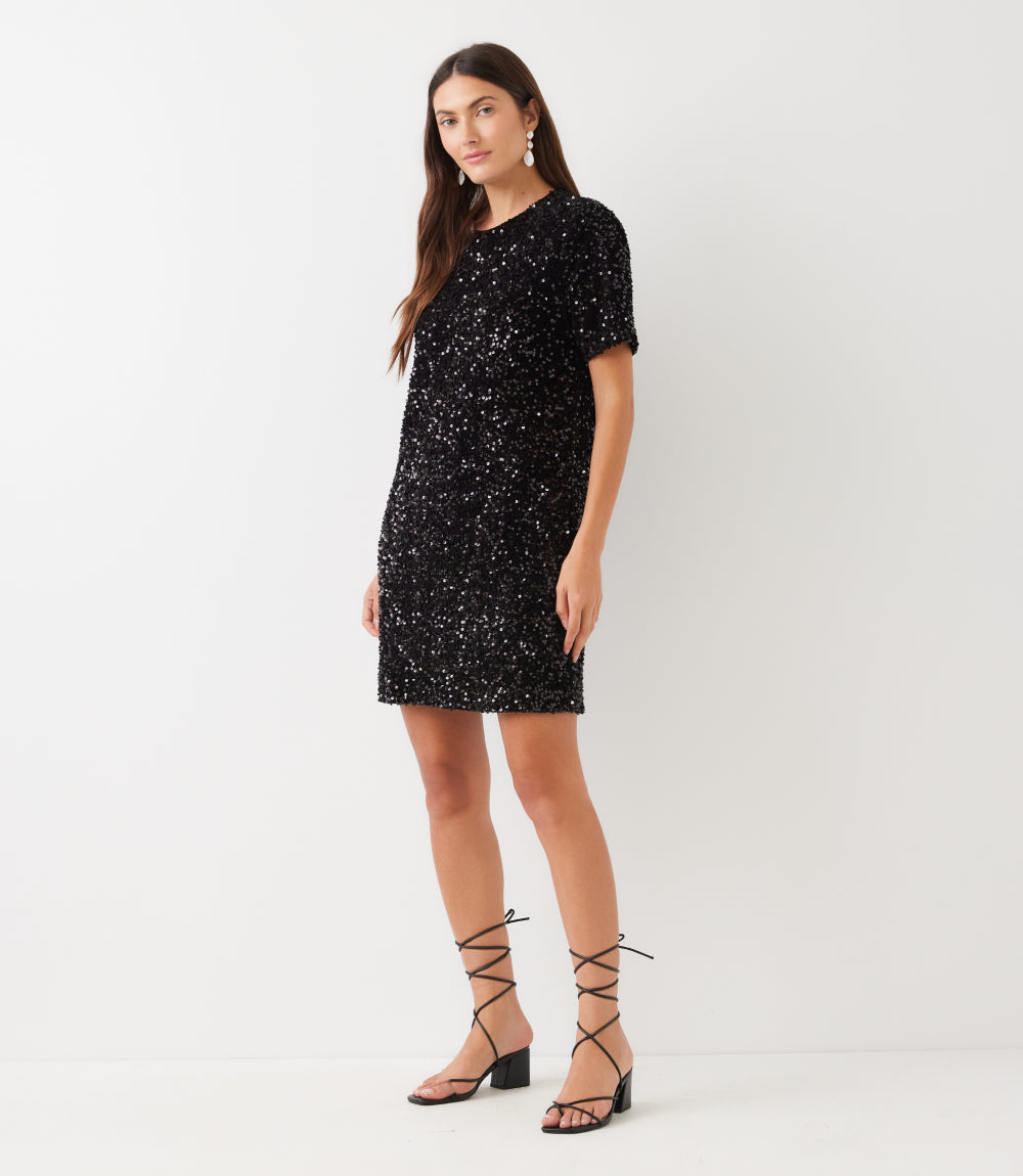 A woman modeling a short-sleeve black sequined mini dress with a crew neck, paired with black lace-up heeled sandals, against a plain white studio background.