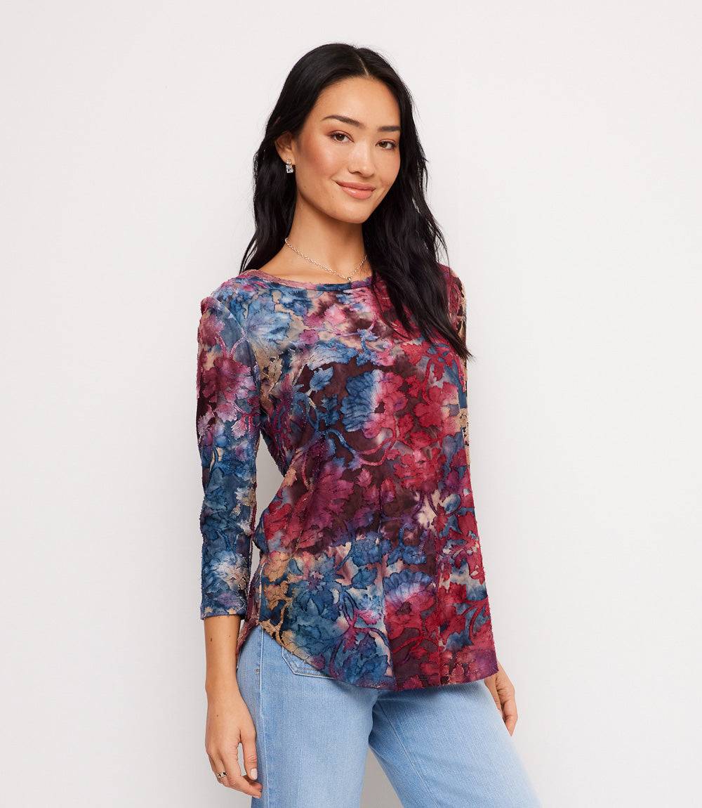 A woman modeling a three-quarter sleeve top with a vibrant blue, red, and purple floral watercolor pattern, paired with light blue jeans against a plain white background.