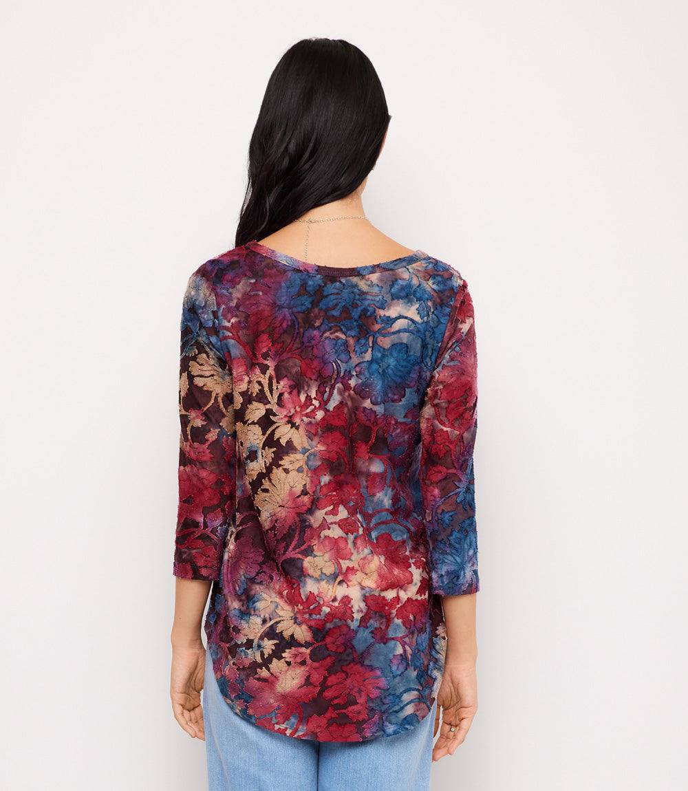 A woman viewed from behind wearing a three-quarter sleeve top with a dense abstract floral pattern in red, blue, and beige, paired with light blue jeans against a plain white background.