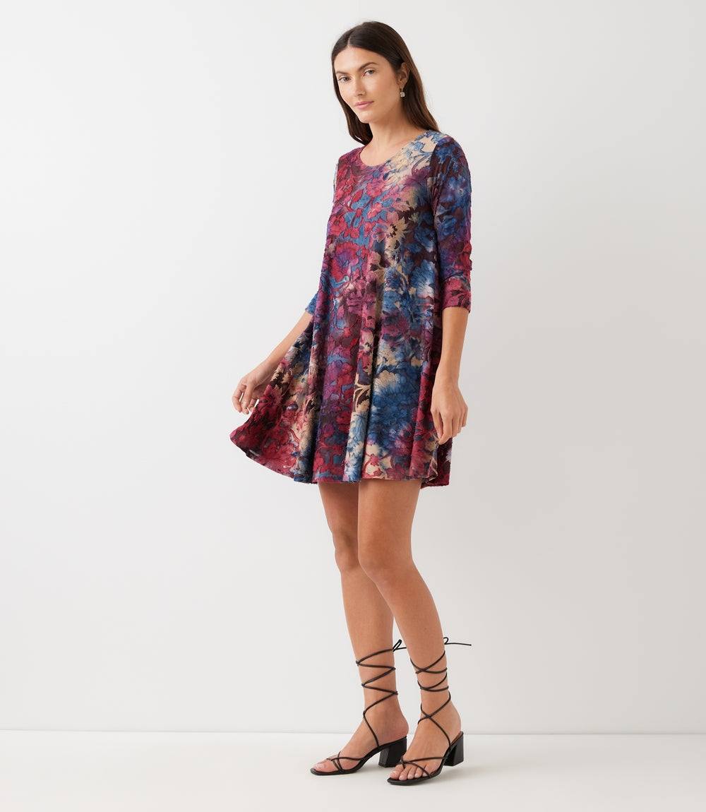 A woman modeling a short, knee-length shift dress with three-quarter sleeves and a vibrant abstract floral print in shades of blue, purple, and red. She is wearing black lace-up flat sandals against a plain white studio background.