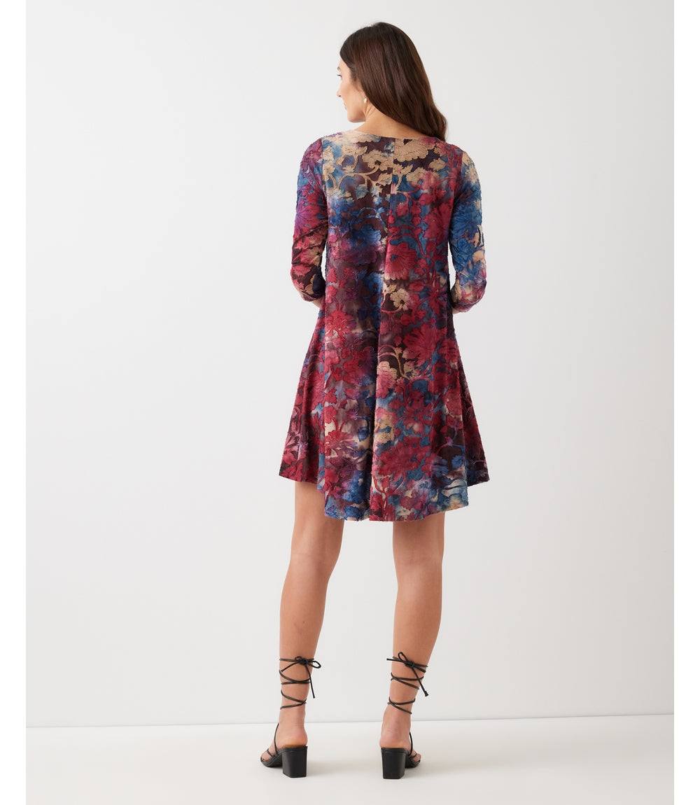A woman viewed from behind wearing a short, long-sleeved floral dress with a red, blue, and purple pattern, paired with black lace-up heels, against a plain light gray studio background.