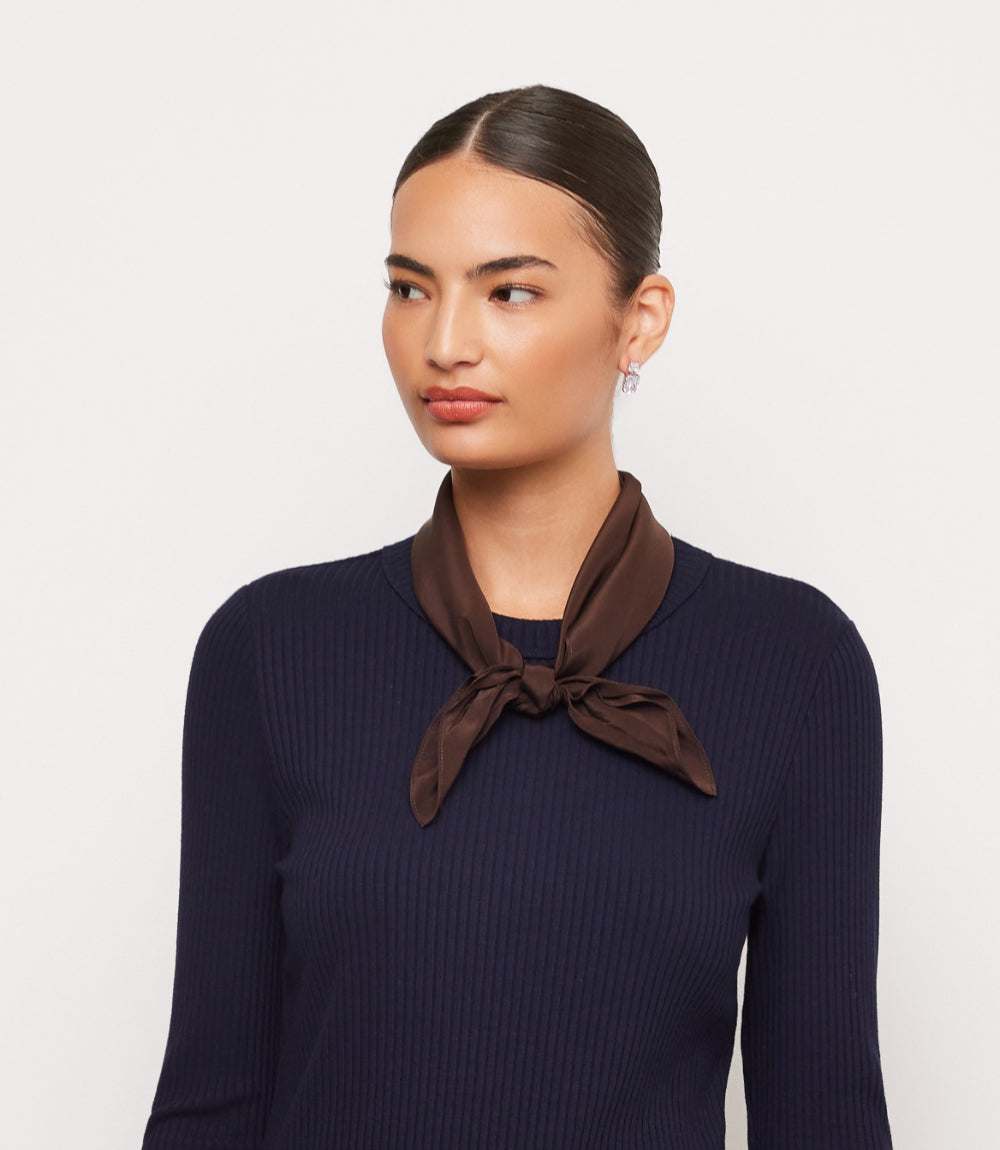 A woman with her hair pulled back wears a dark brown satin neck scarf tied in a knot at the collar of a dark navy ribbed long-sleeve top, photographed against a light gray studio background.
